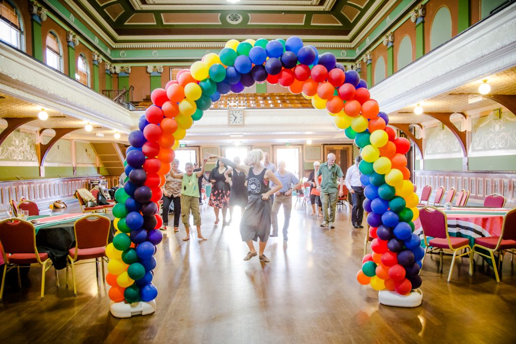 An archway of rainbow balloons is in the foreground of the picture. Behind it are numerous people dancing. They are in a beautiful old hall with a shiny wooden floor, walls painted in green and orange. Tables and chairs line the walls