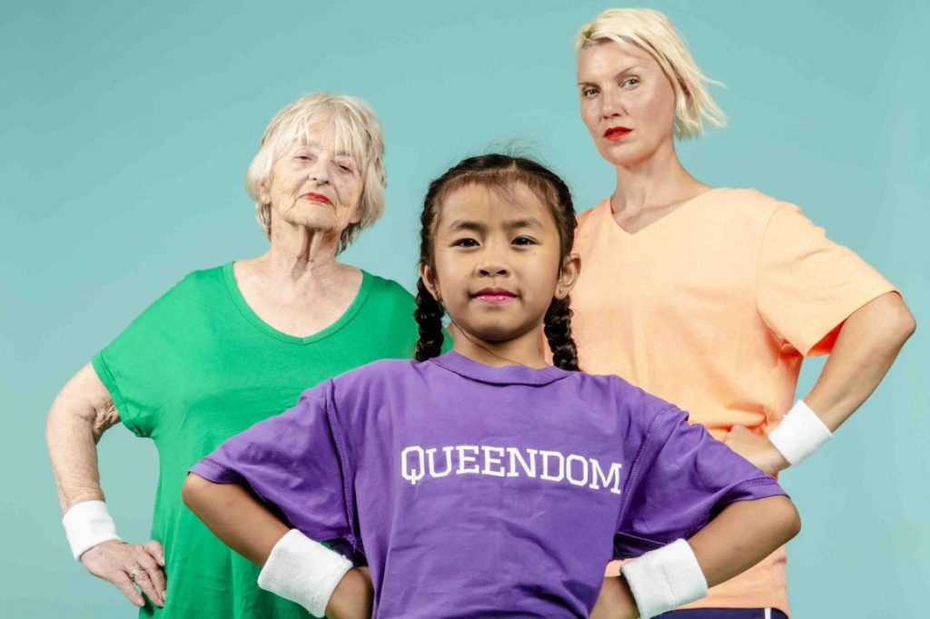 A mid body image of three people standing in front of a light blue background. There is a child in the middle looking at the camera and wearing a shirt with the word 'Queendom' on it.