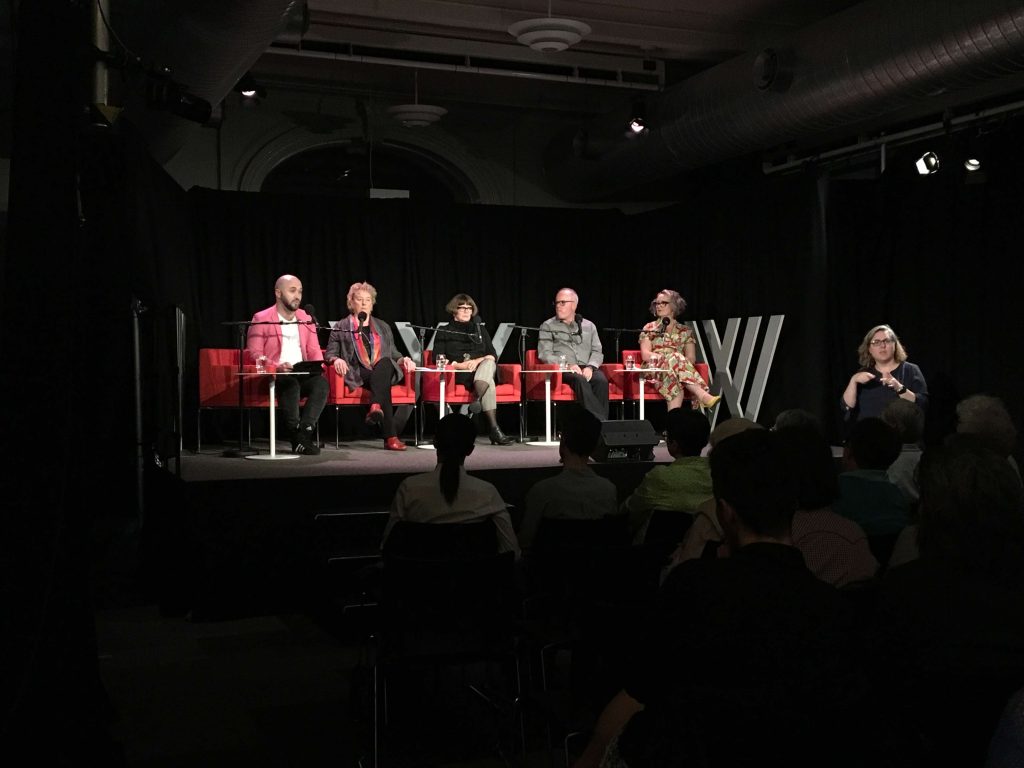 An image of five people wasted and having a conversation on a lit stage. They are all speaking into microphones and an Auslan interpreter in black and wearing glasses is standing in front of the stage.