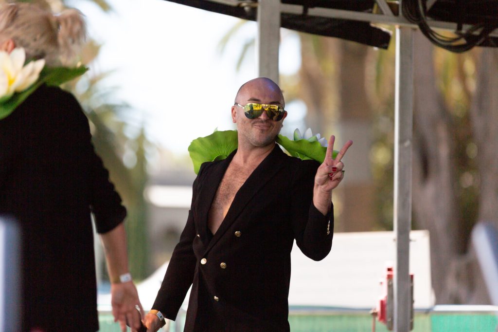 An image of a performer on stage giving a peace sign to the camera. They are wearing a black blazer with large lotus flowers shoulder pads and gold sunglasses.