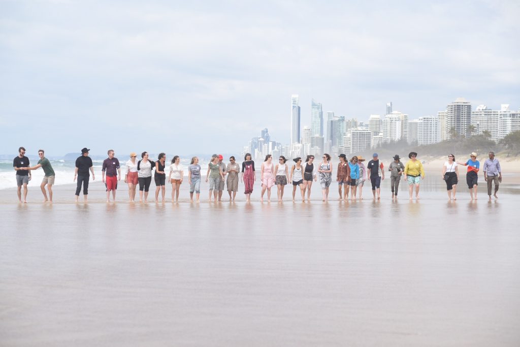 An image of a large line up people all walking down and open stretch of beach with many skyscrapers in the background.