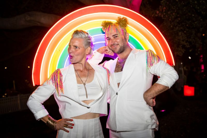 An image of two people wearing white suits and standing in front of a neon rainbow. Their suits have rainbow tassels.