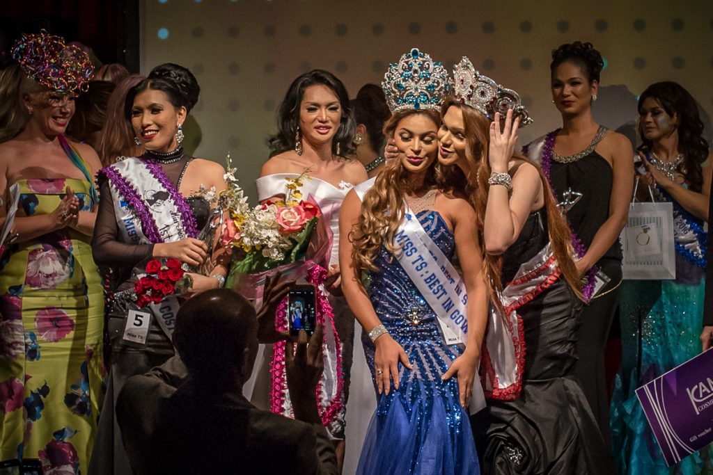 An image of several contestants in a beauty pageants on stage congratulating the crowned winners. They are wearing bright gowns, sashes and holding flowers.