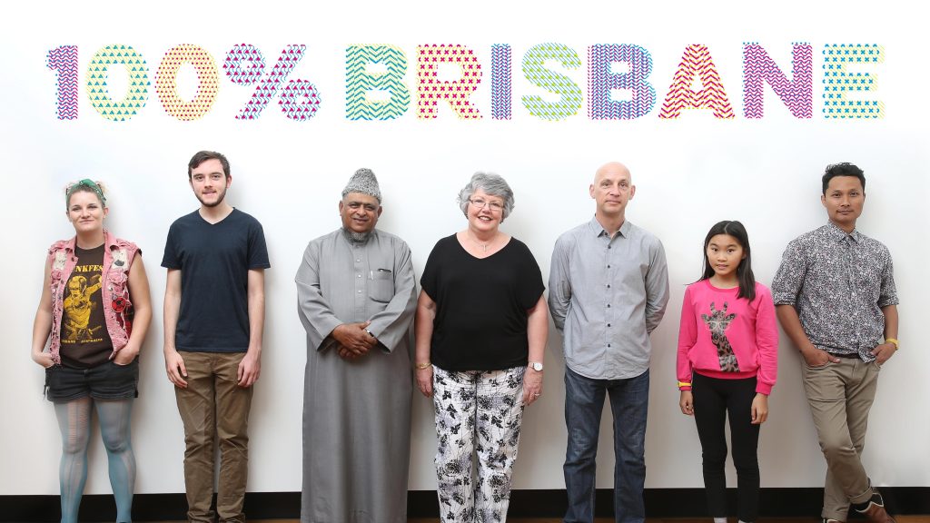 An image of seven people standing in front of a white wall with the words 100% Brisbane on the wall above their heads. They are all looking at the camera.