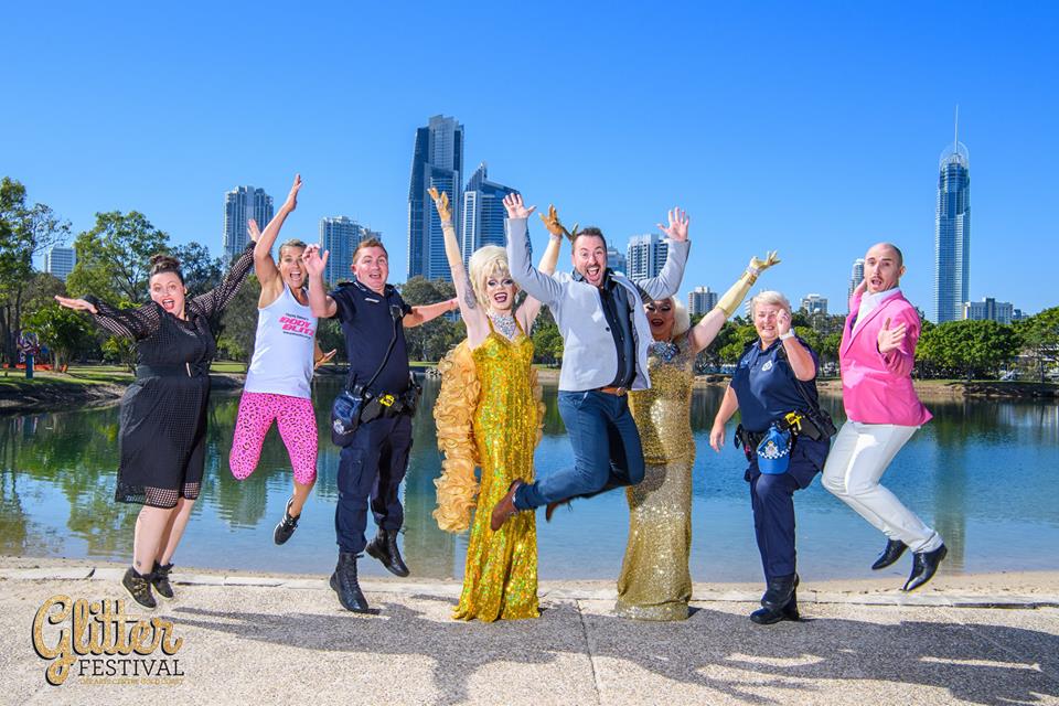 A promotional image of Glitter festival featuring seven people jumping for joy by a tropical waters edge under a blue sky.