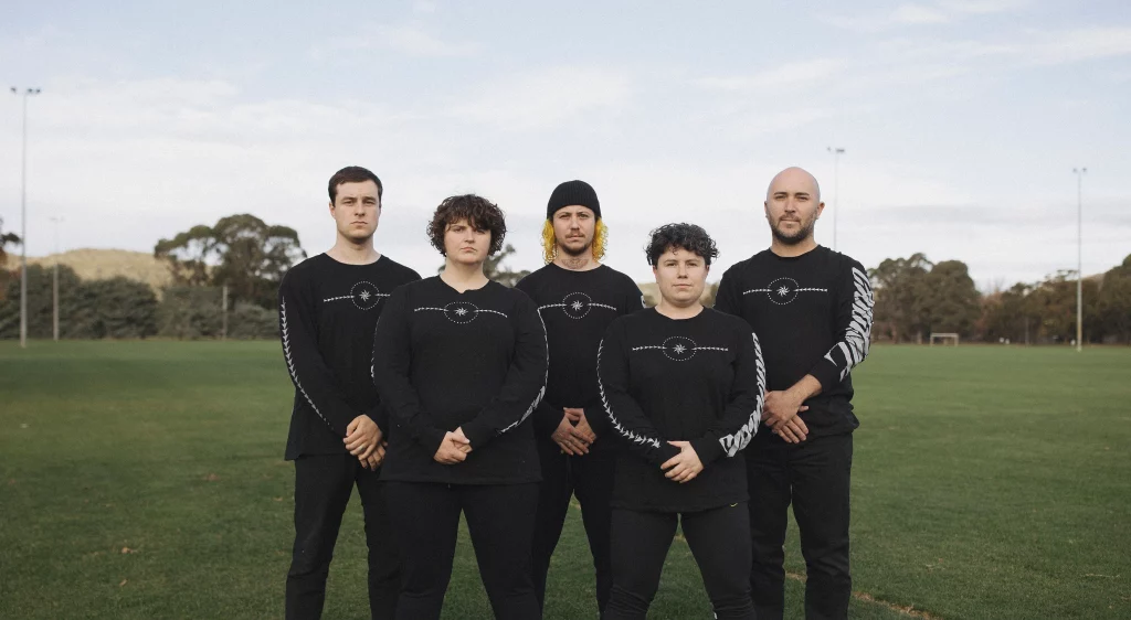 An image of five people standing on an open green sports field all looking at the camera and clasping their hands in front of their bodies. They are all wearing black and white uniforms.