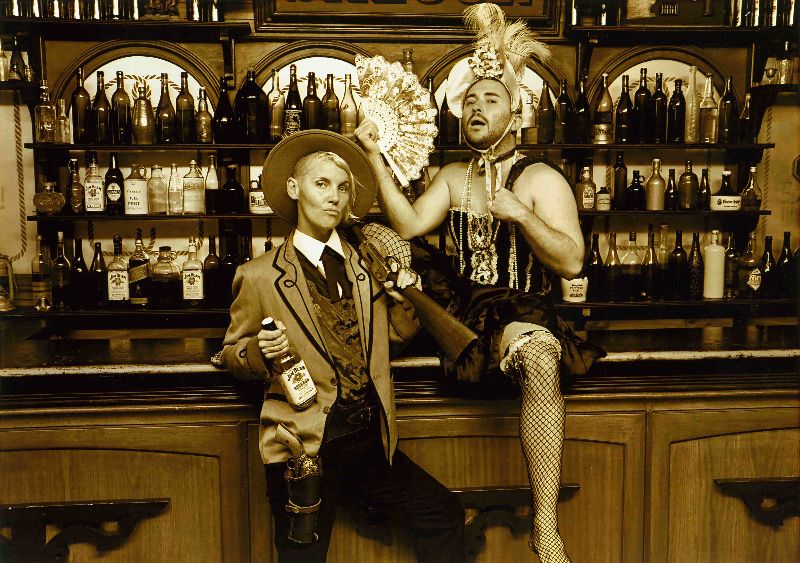 Two people in costume dress from an American Western Saloon. One person is standing at the bar with whisky pretending to be a gunslinger. The other person is sitting on the bar in can can dress and holding a fan.