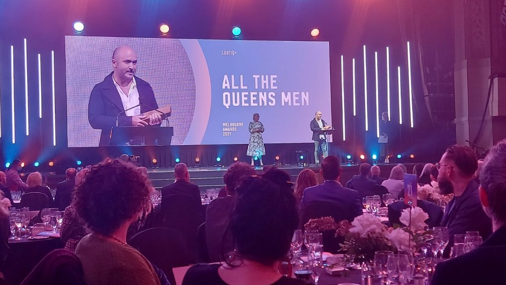 An image of an awards ceremony featuring a solo caucasian male on a large screen and the same person at a podium holding an award and giving a thank you speech.